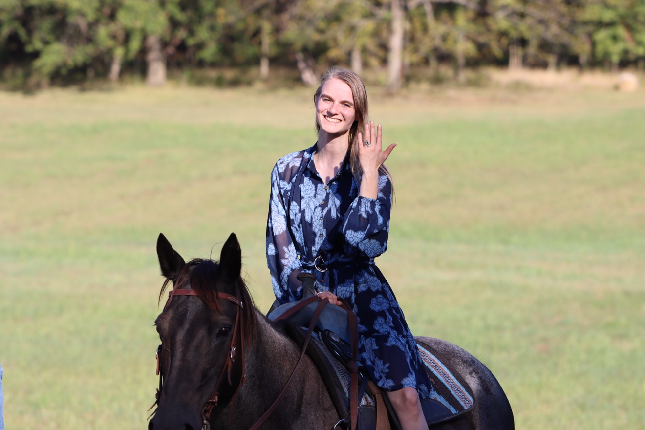 Kristi sitting on her horse, smiling and holding up her hand to show off the engagement ring